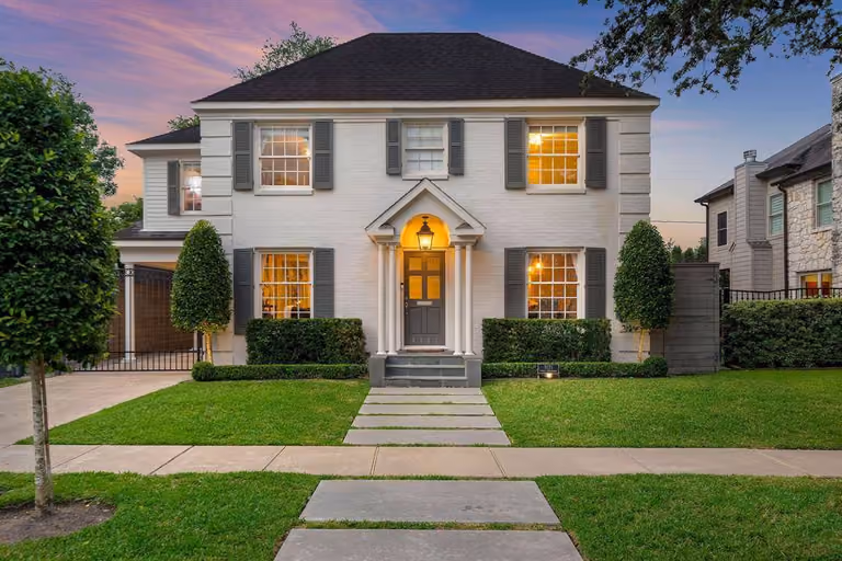 Dusk at a two-story West University colonial: white-painted brick behind a columned front portico, a lantern glowing over the door, shuttered windows lit warm, and a stepping-stone walk crossing the front lawn.